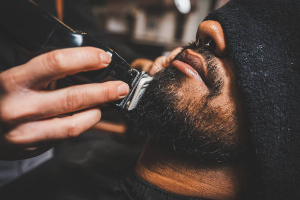 A Caucasian hand with an electric cutting machine passes over the beard of an African-American Brazilian boy to fix it. It is a barbershop, the client has his eyes covered with a towel. the photo is dark and vintage or hipster