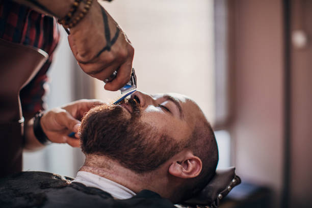 Young bearded man getting beard haircut by young male hairdresser while lying in chair in a barber shop.